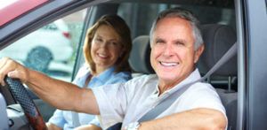 Smiling, happy older couple driving a car together, with the man wearing a seatbelt and looking at the camera.
