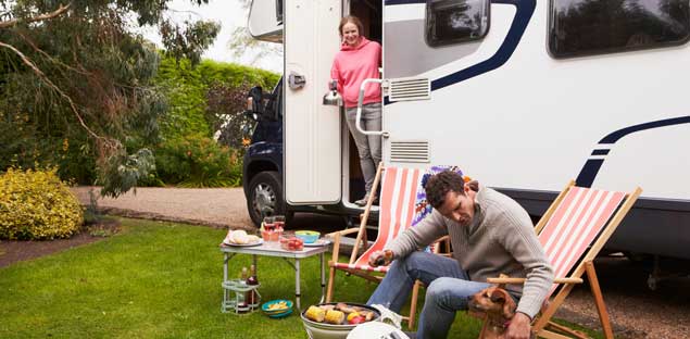 A couple enjoying a barbecue and relaxing with their dog outside a parked RV or motorhome at a campsite.